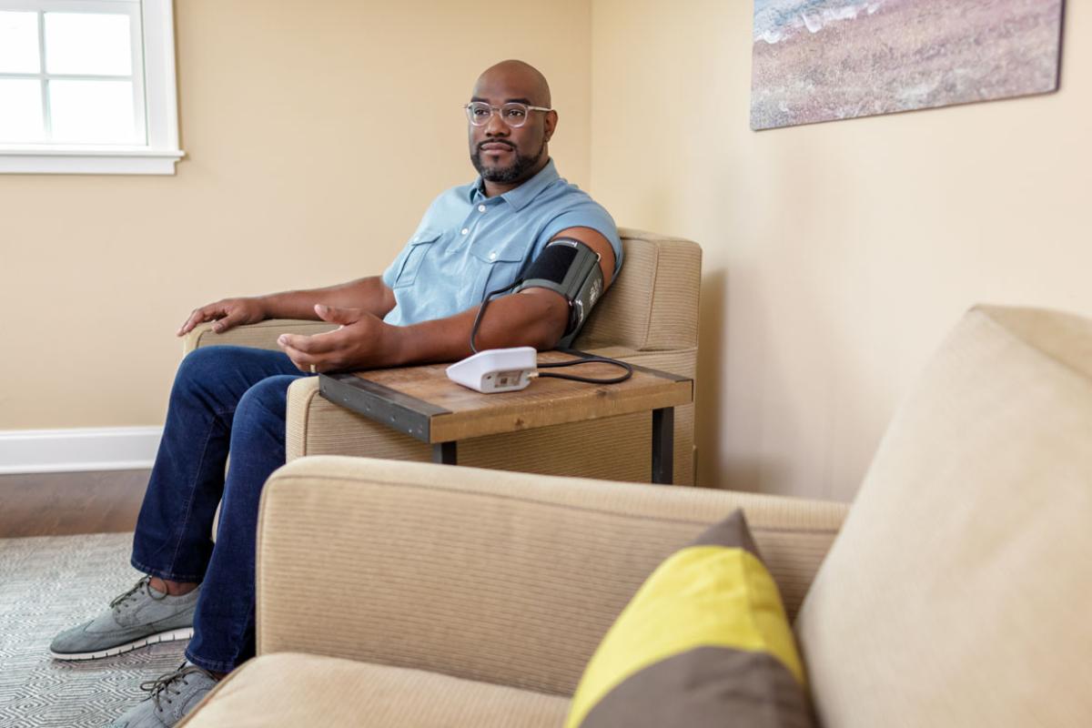 African-American man sitting in a chair, taking his blood pressure using a portable monitor.