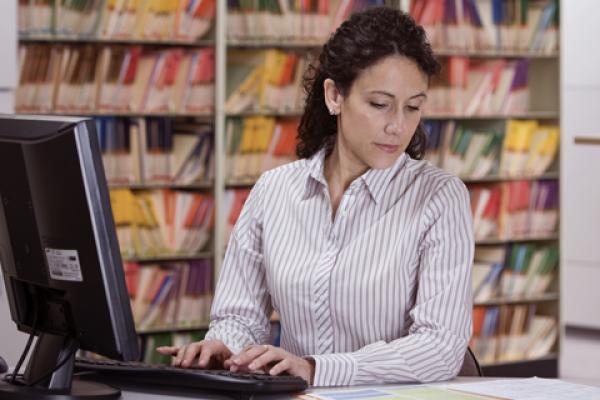 A physician works at a PC while looking down at paperwork.